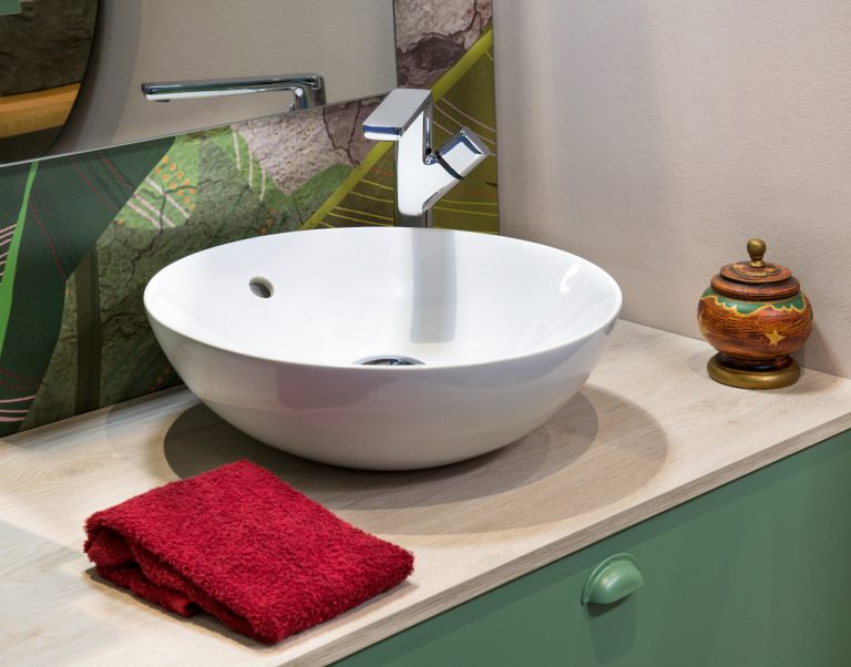 Interior of bathroom with reflecting mirror fixed on green design tiles wall while folded red towel placed near modern white wash basin with tap and incense burner vase in light